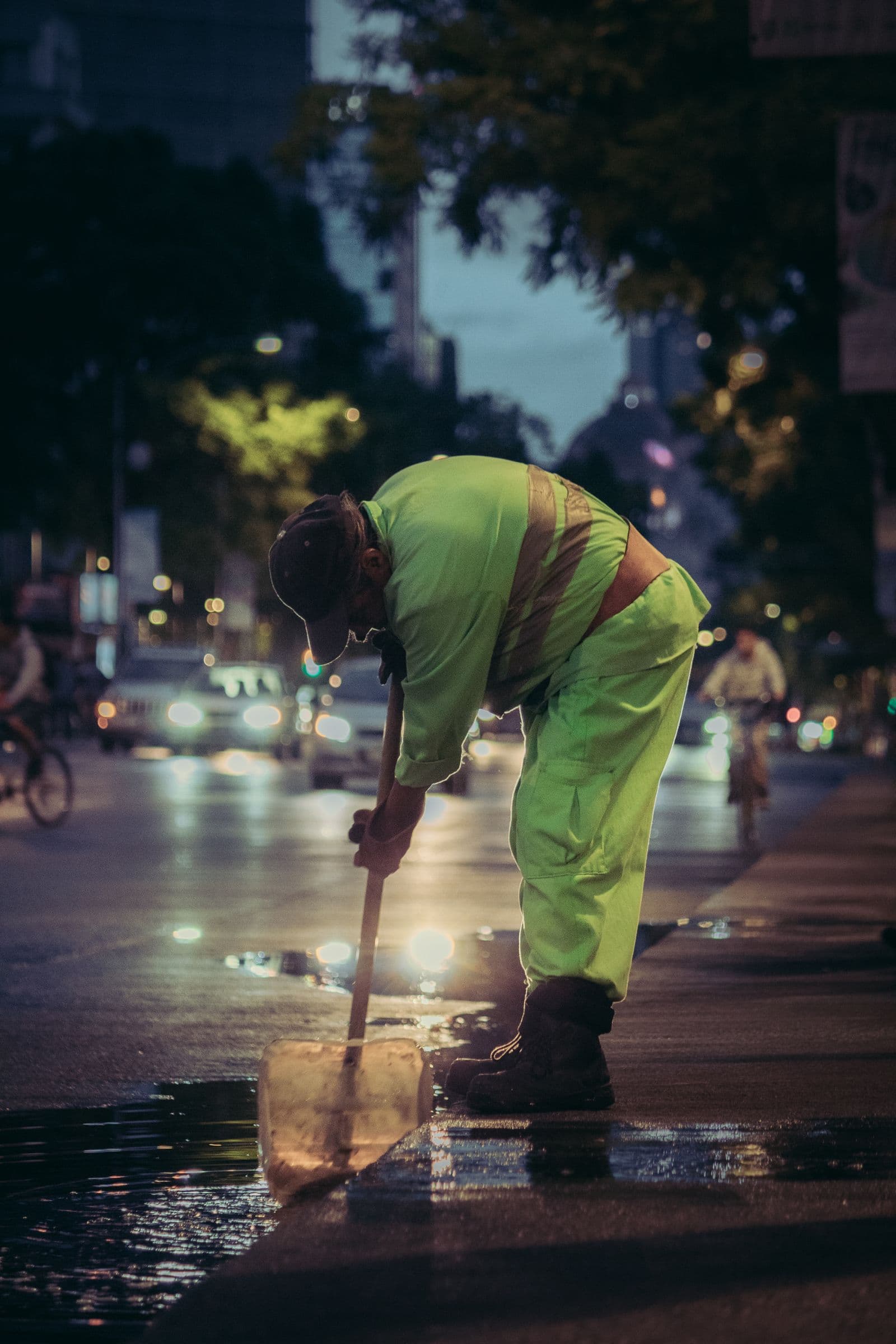 Trabajador de limpieza viaria con ropa amarilla de alta visibilidad y bandas retrorreflectantes barriendo una calle mojada al anochecer, con faros de coches al fondo