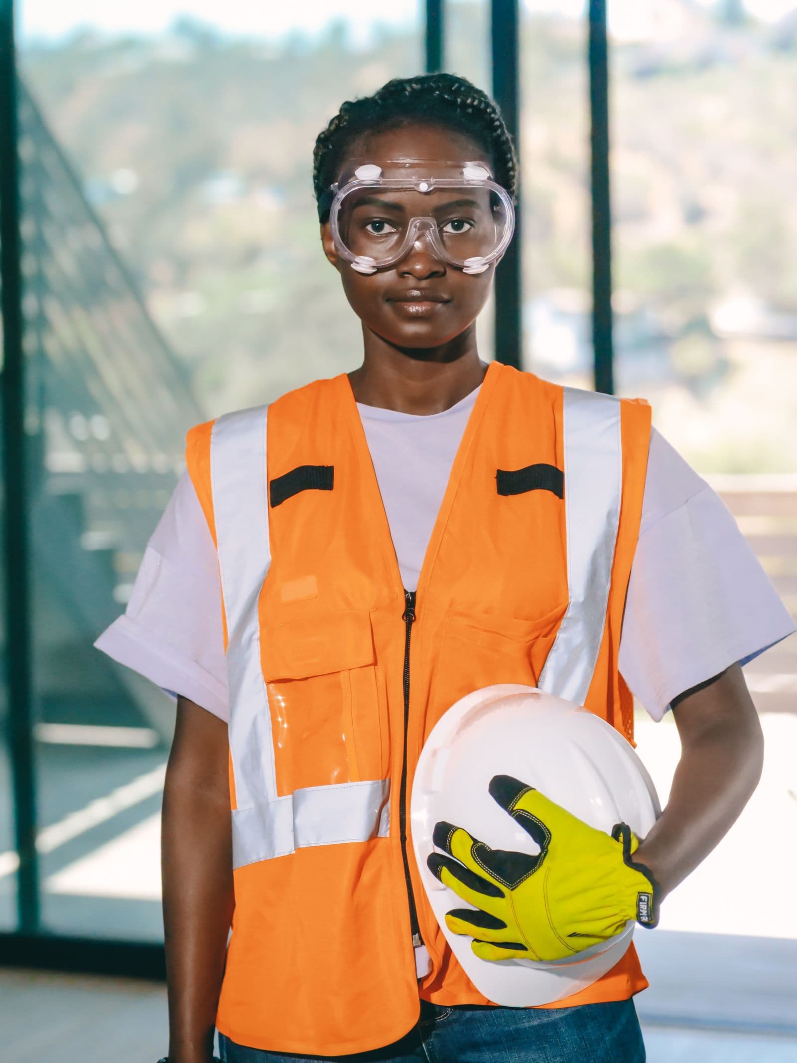 Trabajadora con chaleco naranja de alta visibilidad con bandas retrorreflectantes, gafas de protección y un casco blanco en la mano